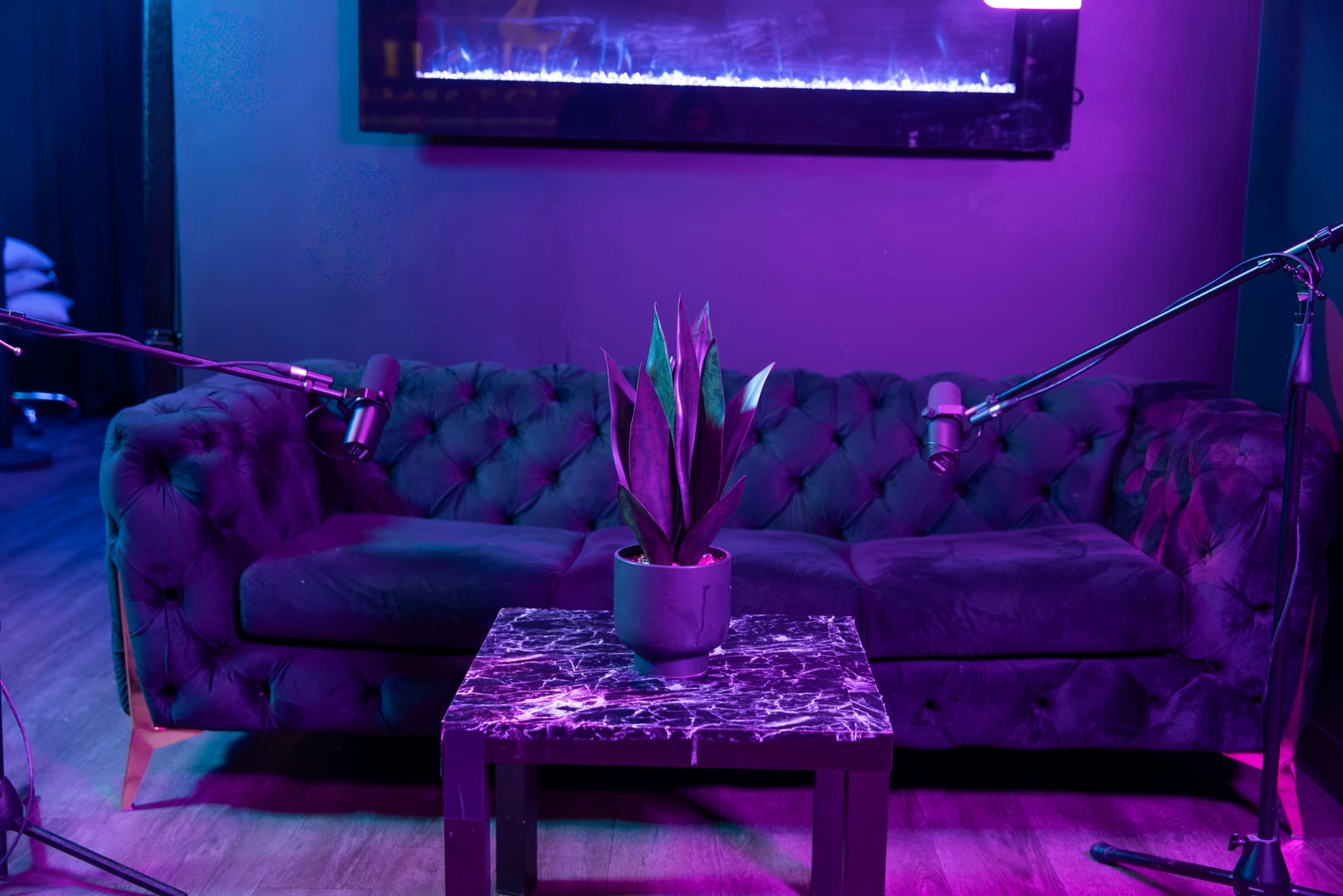 The Fireside set at Lush Event Space — tufted velvet sofa, two broadcast microphones on booms, wall fireplace glowing above, marble table, violet light